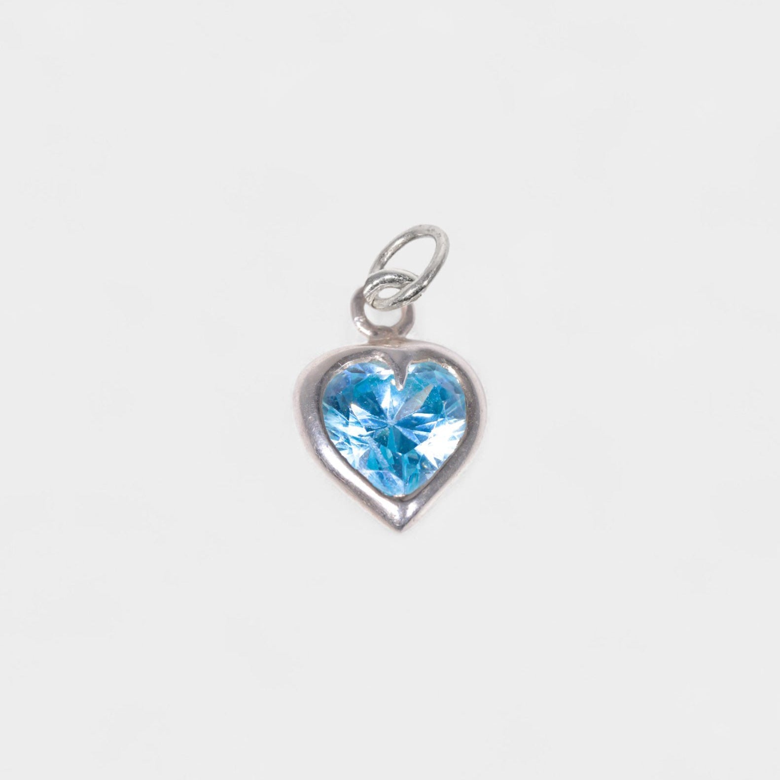 A silver heart-shaped charm featuring a faceted light blue gemstone, shown against a plain light gray background.