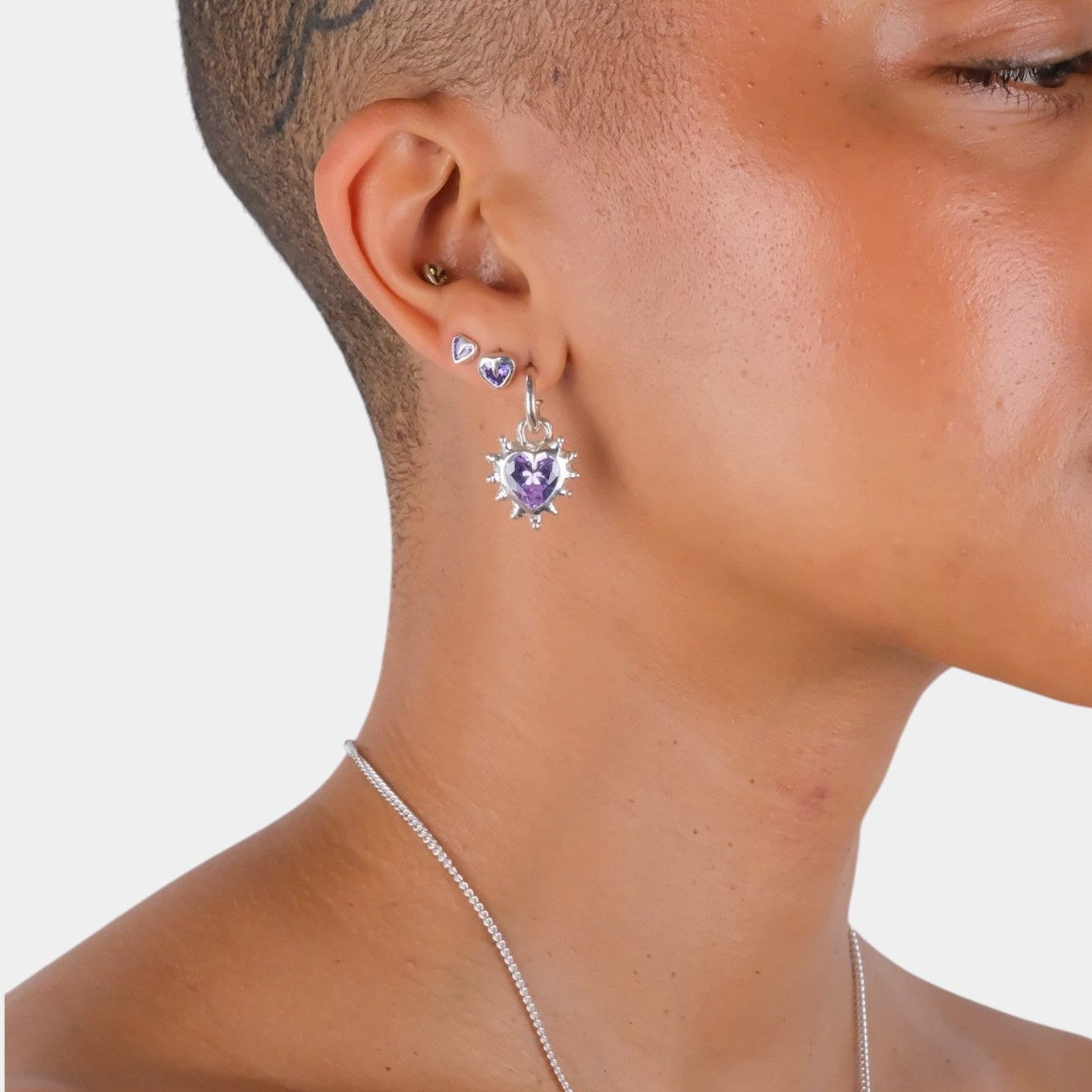 Close-up of a person wearing purple heart-shaped earrings on a plain background