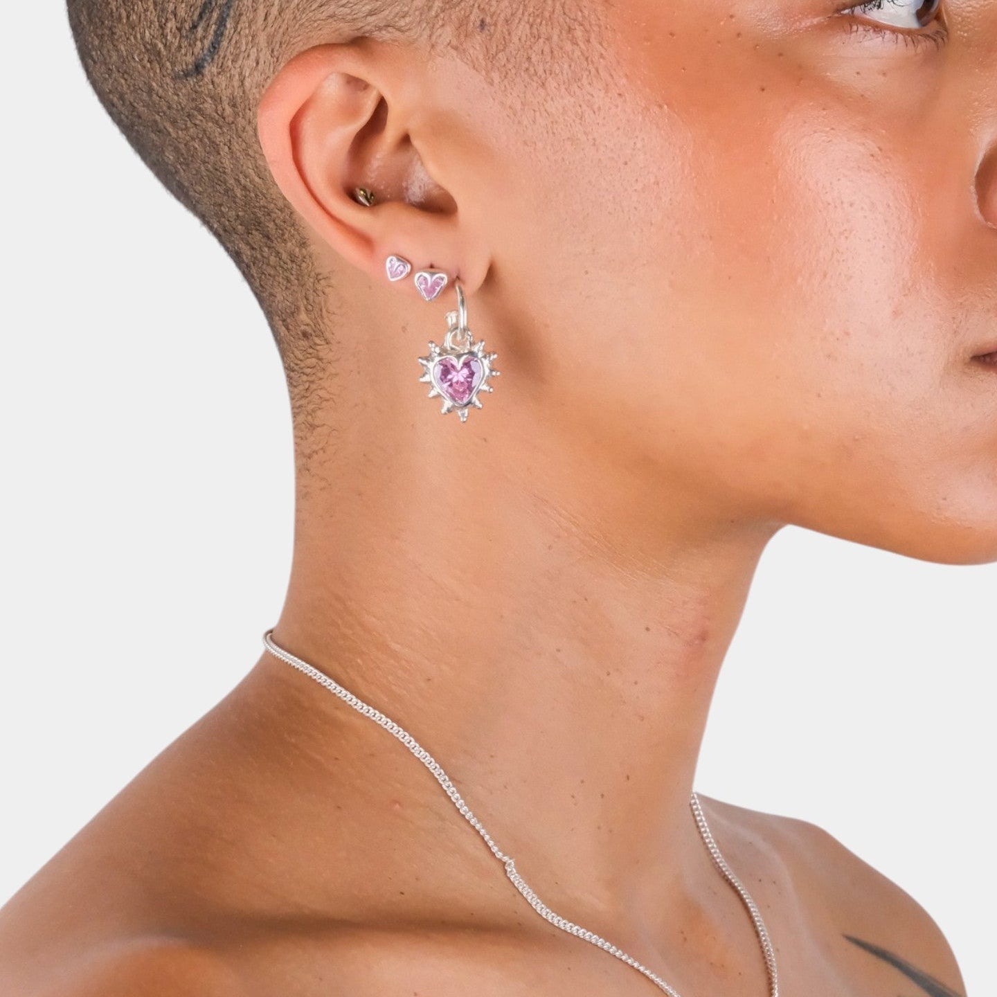 Close-up of a person wearing heart-shaped earrings with pink gemstones on a plain background