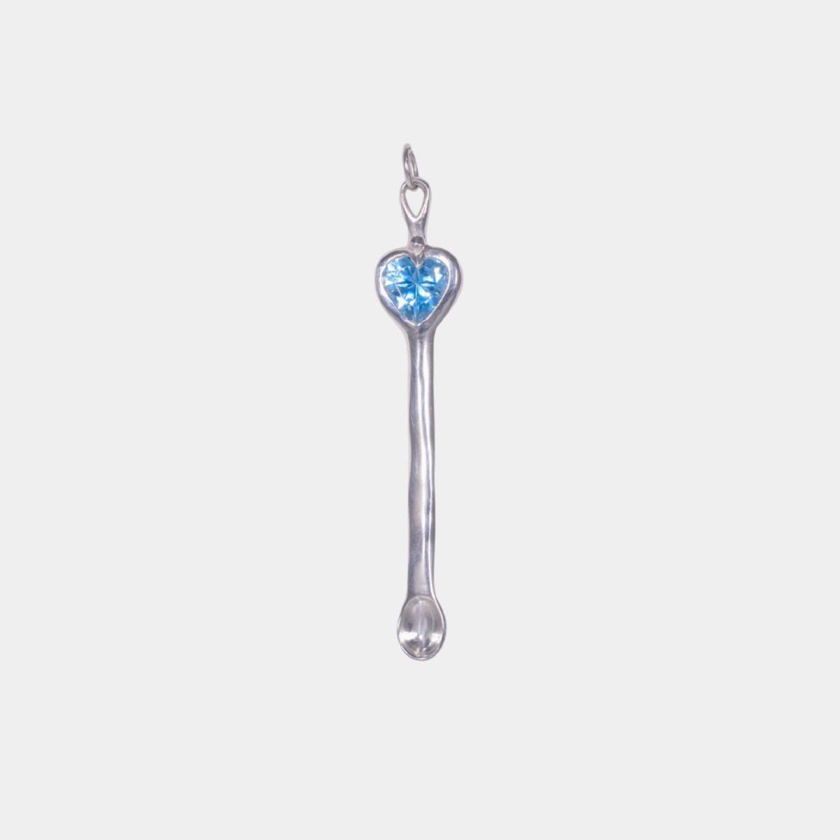 A silver spoon charm pendant featuring a small heart-shaped light blue gemstone at the bowl, set against a plain light gray background.
