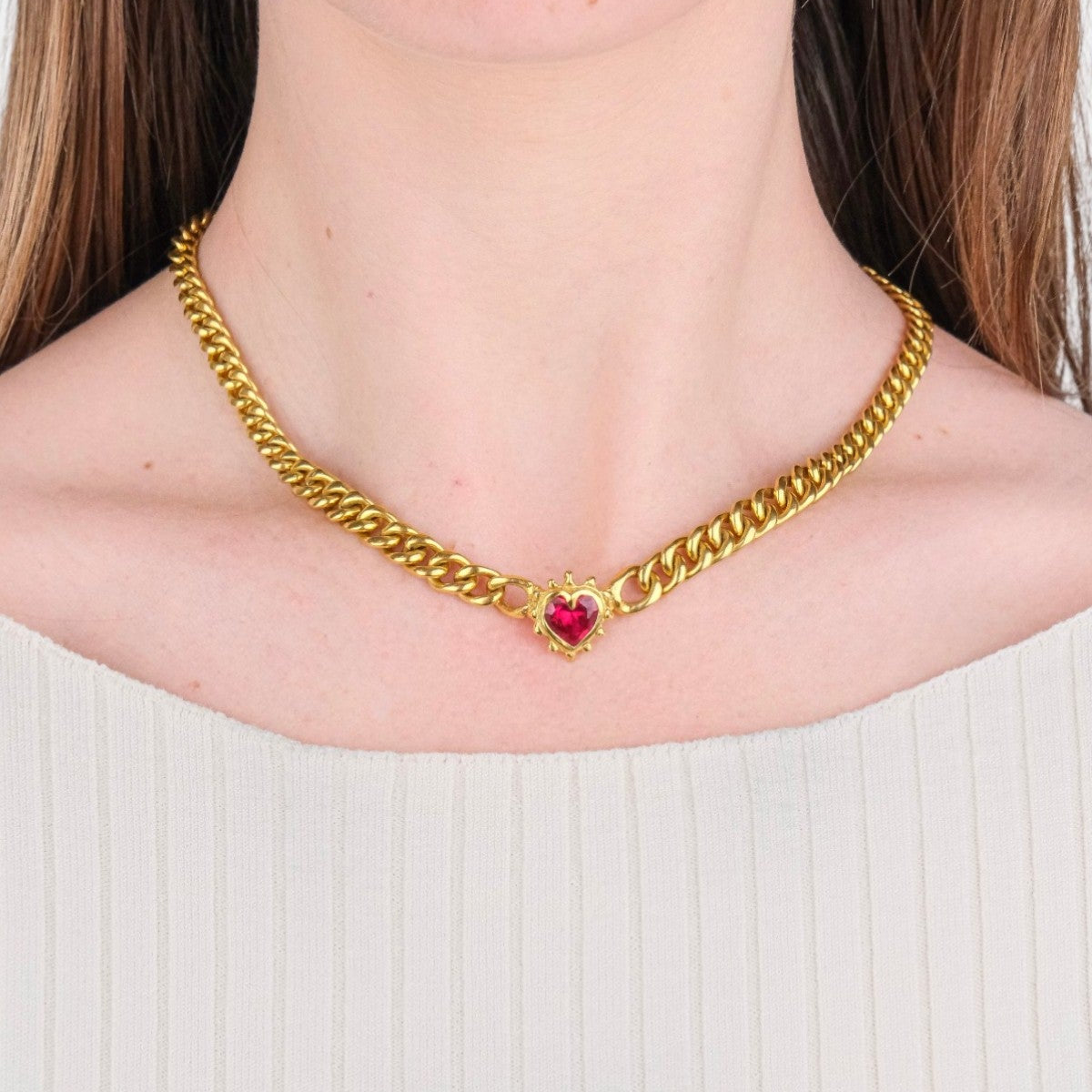 A close-up of a model wearing a gold curb chain necklace with a small red heart charm at the center.