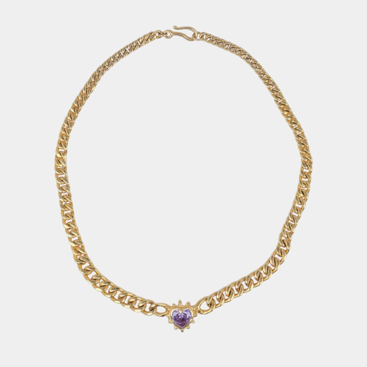 A gold-toned curb chain necklace with a small heart-shaped purple gemstone pendant, displayed on a light gray background.