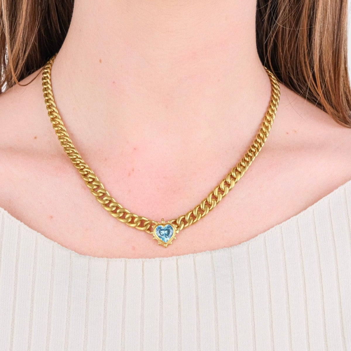 A close-up of a model wearing a gold curb chain necklace with a small heart-shaped blue gemstone pendant.