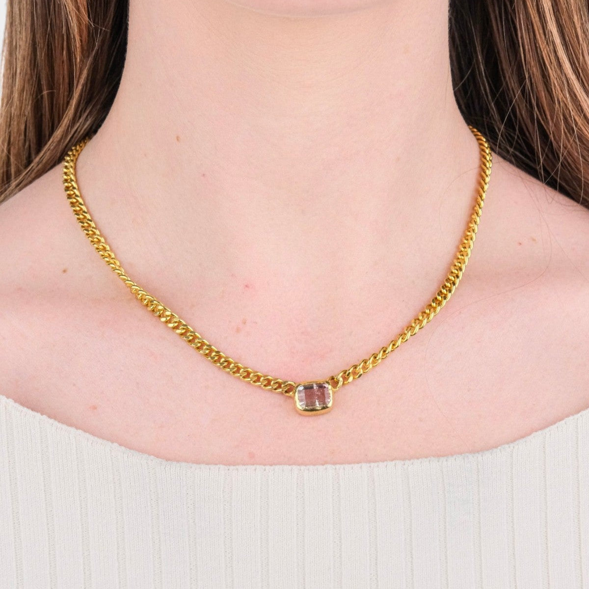 A close-up of a model's neck and lower face wearing a gold chain necklace with a small rectangular pendant featuring a pinkish stone.