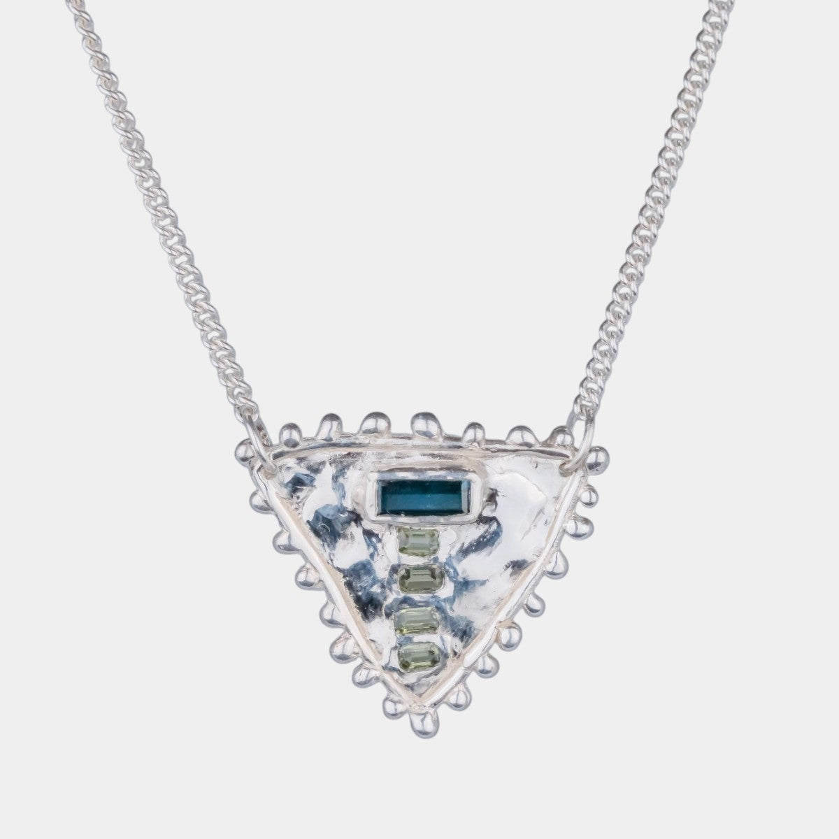 A silver triangle pendant necklace featuring a textured silver center with a small dark blue rectangular stone and a vertical row of clear stones, suspended on a delicate silver chain against a light gray background.
