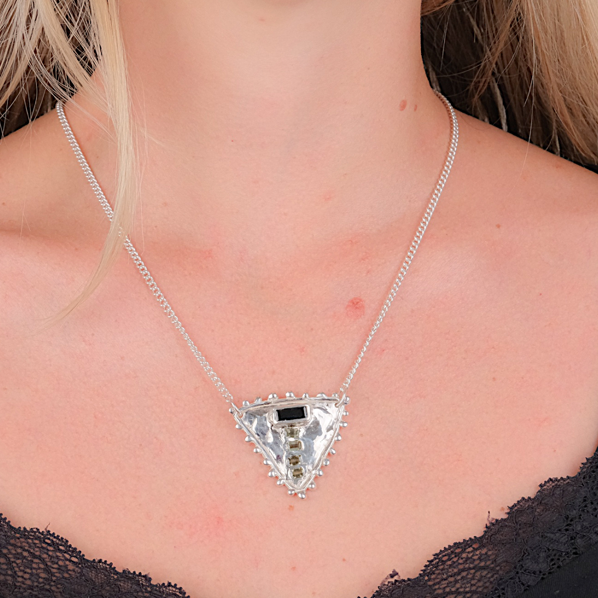 A close-up of a woman wearing a silver triangular pendant necklace with a clear faceted center stone and a small black rectangular accent, paired with a black lace-trim top.