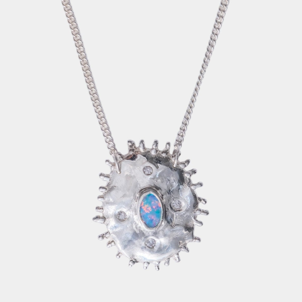 A silver chain necklace with a textured, organic-shaped silver pendant featuring a central blue opal-like inlay, photographed against a light gray background.