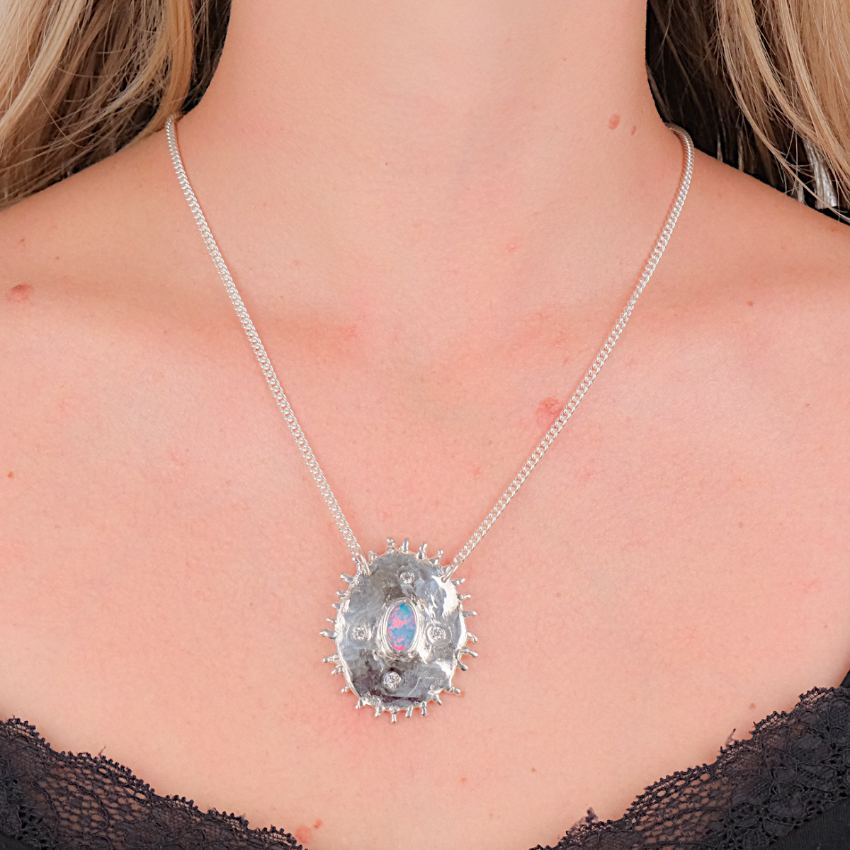 A close-up of a model wearing a silver chain necklace with a round, textured silver pendant featuring a central iridescent blue and pink stone.