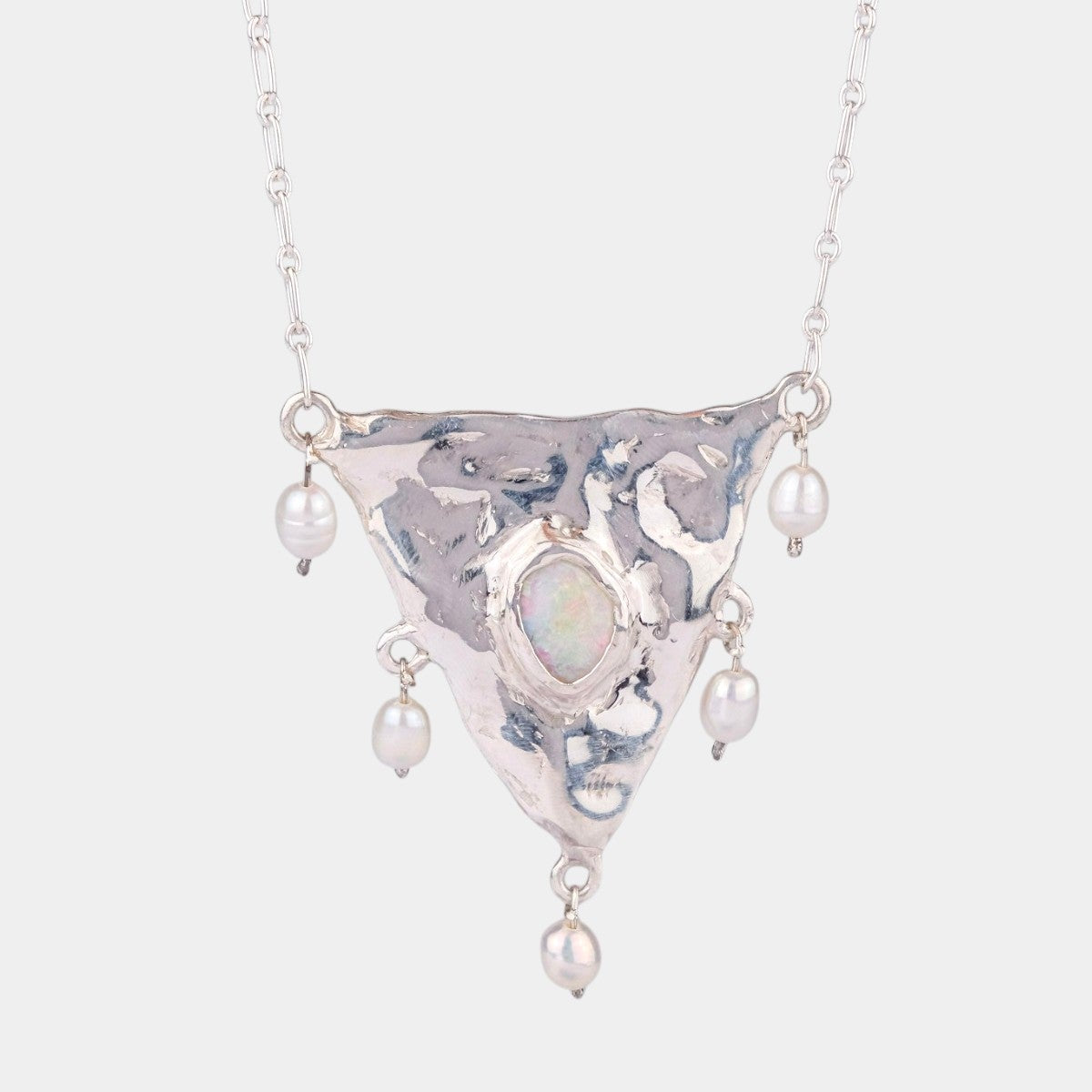A silver-toned necklace featuring a hammered triangular pendant with a central iridescent opal-like stone and dangling pearl beads, shown against a light gray background.