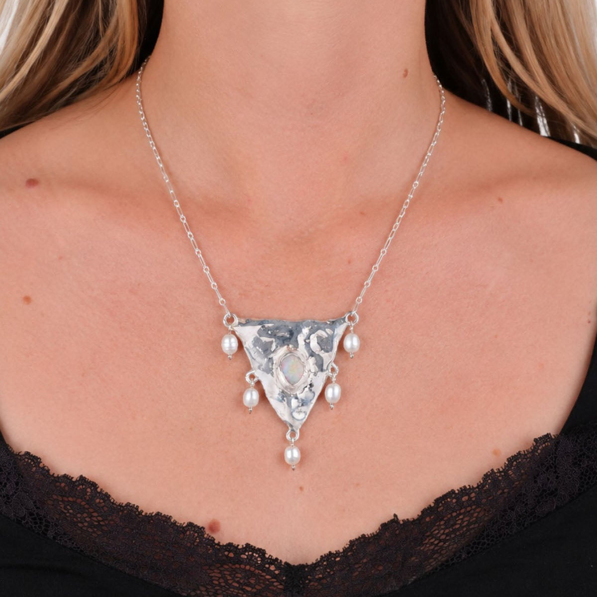 A close-up of a woman wearing a silver triangular pendant necklace with dangling pearl-like beads, paired with a black lace-trim top.
