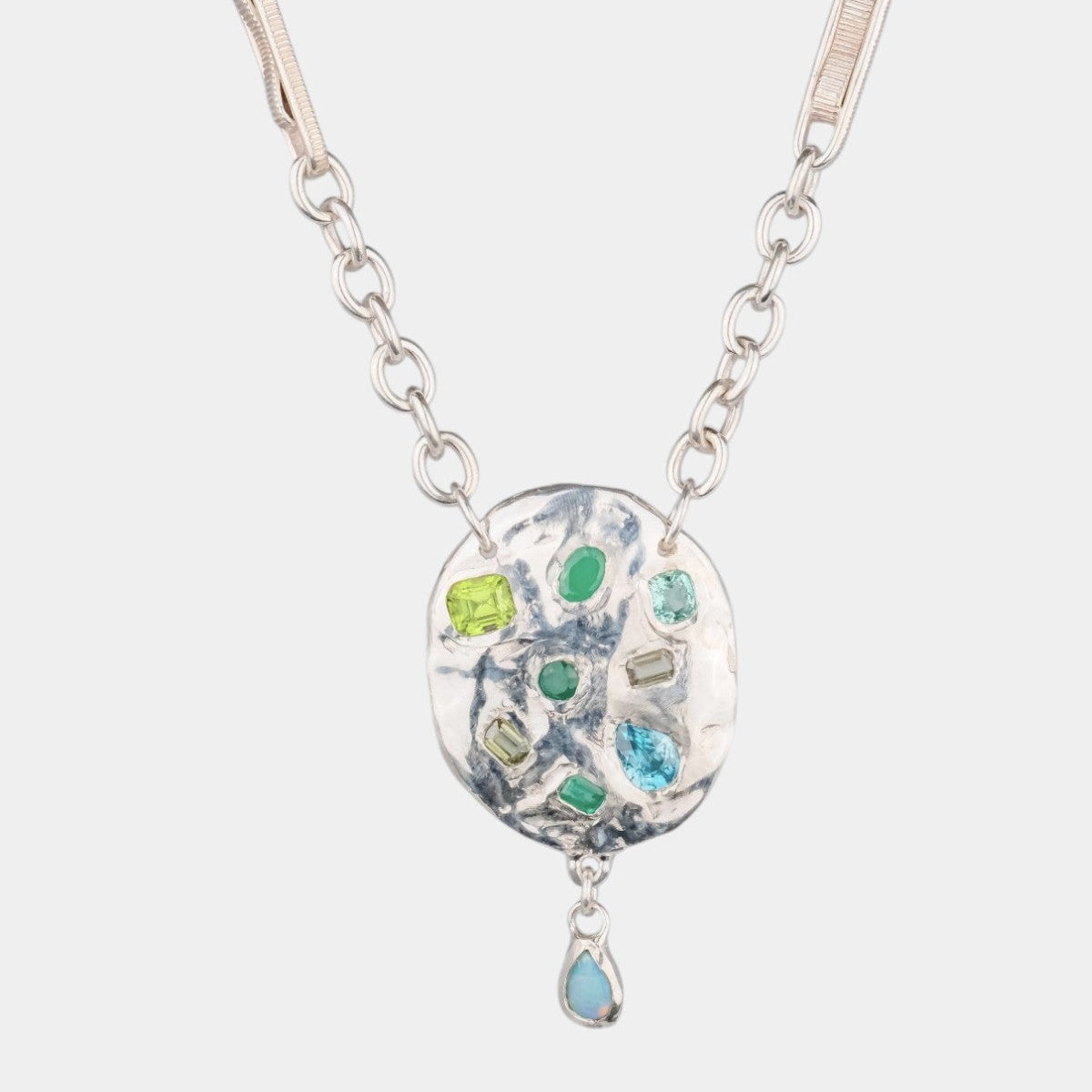 A silver-tone necklace featuring a textured, irregularly shaped pendant with embedded green and blue gemstones, and a small teardrop-shaped blue stone hanging below, shown against a light gray background.