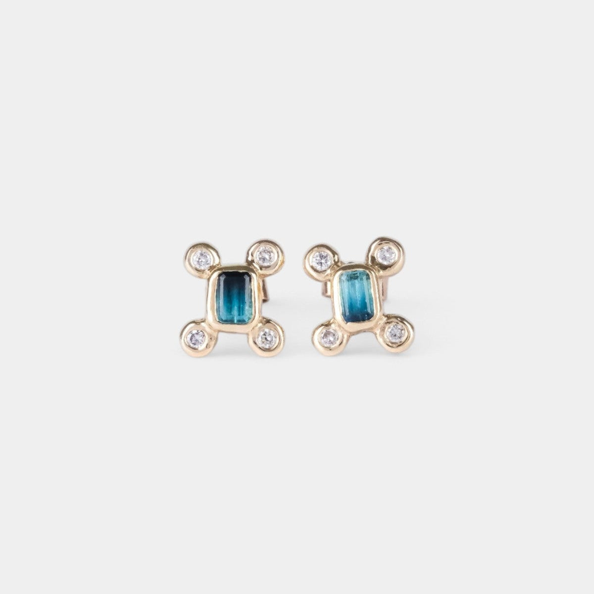 A pair of gold-toned stud earrings featuring a central rectangular blue stone surrounded by four small clear round stones, set against a plain light gray background.