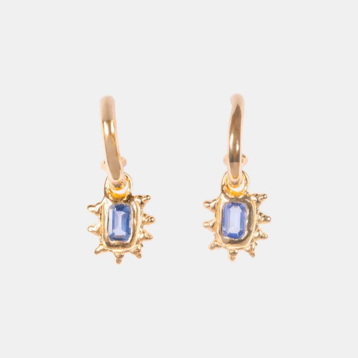 A pair of gold huggie hoop earrings with a small sunburst halo design and a rectangular blue gemstone in the center, shown on a light gray background.