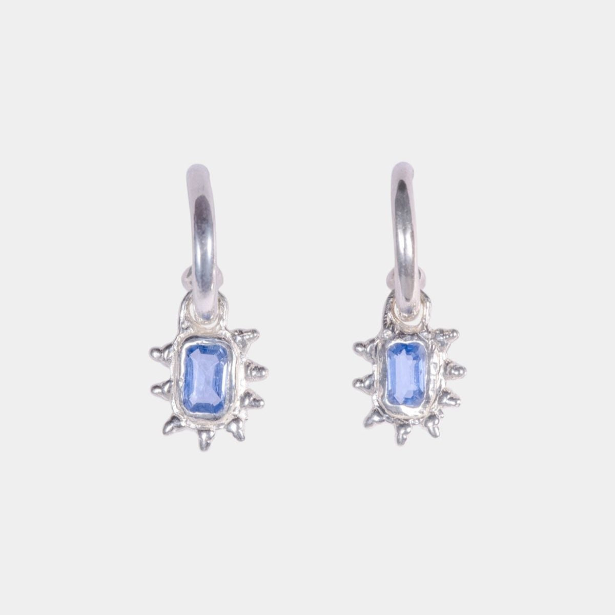 A pair of silver dangle earrings featuring a rectangular blue gemstone in a sunburst setting, shown against a plain light gray background.